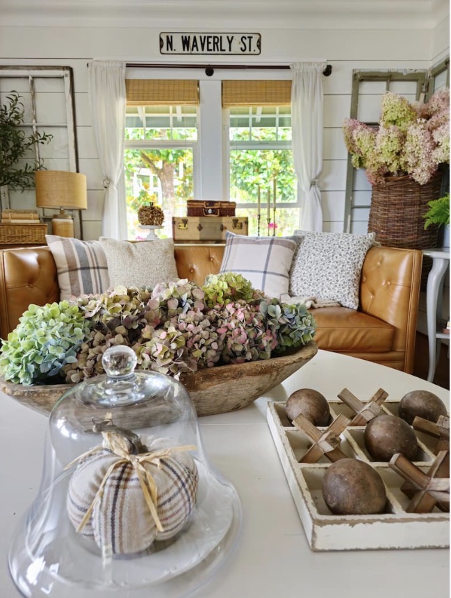 Cozy living area featuring a brown leather couch, decorative throw pillows, and a wooden coffee table adorned with a glass cloche holding a plaid pumpkin and a rustic bowl filled with dried hydrangea flowers. Natural light filters through the windows with white curtains, and decorative items include a vintage suitcase and potted plants.